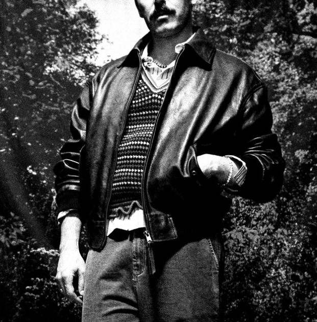 Black and white portrait of Sam Palladio wearing an open brown leather jacket over a chunky knit sweater, standing outdoors in front of foliage